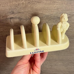 Può includere: Un porta-toast in ceramica con quattro fessure e una piccola statuetta di un uomo sul lato. Il porta-toast è di colore crema e ha la scritta "Lurpak" stampata sul davanti.