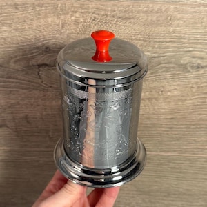 Pode incluir: Um recipiente prateado brilhante com tampa e botão vermelho brilhante. O recipiente tem um design floral em relevo ao redor do corpo. O recipiente é feito de metal e tem um visual decorativo e vintage.