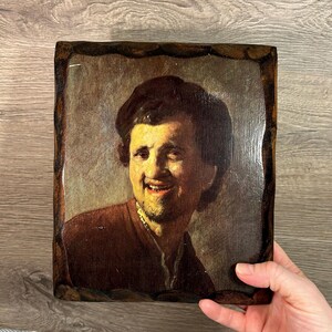 May include: A framed painting of a man with a dark brown beard and moustache, wearing a dark brown jacket and a black hat. He is smiling and looking to the left. The painting is on a wooden frame.