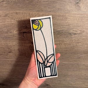 May include: A rectangular ceramic tile featuring a stylised floral design with a yellow flower head, pink leaves, and black outlines on a light grey background. The artwork is framed by a thick black border with vertical lines at the base.