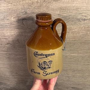 Könnte beinhalten: Ein brauner Keramikkanister mit Henkel und Korken. Auf dem Kanister stehen die Worte "Countryman Farm Scrumpy" und eine Illustration eines Pferdegespanns. Der Krug wird von einer Person gehalten.