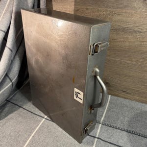 Puede incluir: Una caja de herramientas rectangular de metal gris con asa y pestillo. La caja de herramientas tiene un aspecto ligeramente desgastado y una pegatina en la parte delantera. El asa es curva y el pestillo es un simple cierre metálico.