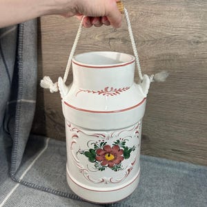 Könnte beinhalten: Weiße Keramik-Milchkanne mit rosafarbenem Blumendesign und einem Seilhenkel. Die Kanne hat ein rustikales, vintage-mäßiges Aussehen.