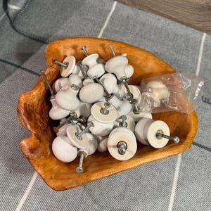 May include: A collection of white ceramic cabinet knobs with metal screws, displayed in a rustic wooden bowl. The knobs have a glossy finish and various shapes. Some knobs are still in a plastic bag. The bowl is made of light brown wood.