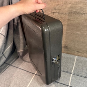 May include: A gray metal briefcase with a handle and a lock. The briefcase is held by a person's hand. The Helix label is visible on the side of the briefcase. The briefcase is sitting on a gray and white plaid blanket.