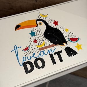 May include: A white rectangular surface featuring a graphic of a toucan with a large orange beak, perched on a brown branch. The design includes small yellow bananas, red stars, blue stars, and watermelon slices, with the text Toucan DO IT.