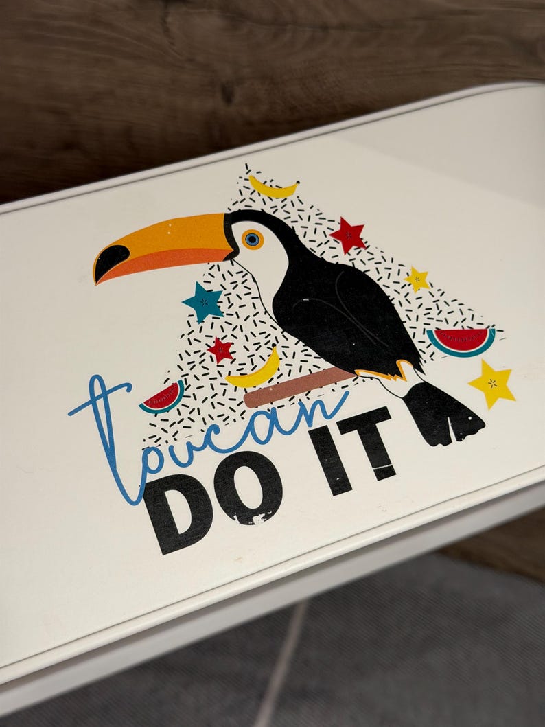 May include: A white rectangular surface featuring a graphic of a toucan with a large orange beak, perched on a brown branch. The design includes small yellow bananas, red stars, blue stars, and watermelon slices, with the text Toucan DO IT.