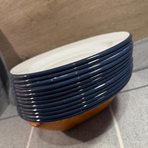 Pode incluir: Uma pilha de doze pratos redondos com uma borda azul-escura brilhante e um centro branco liso. Os pratos est&atilde;o dispostos numa pilha vertical, exibindo as bordas azuis uniformes e o design circular limpo do conjunto de lou&ccedil;as.