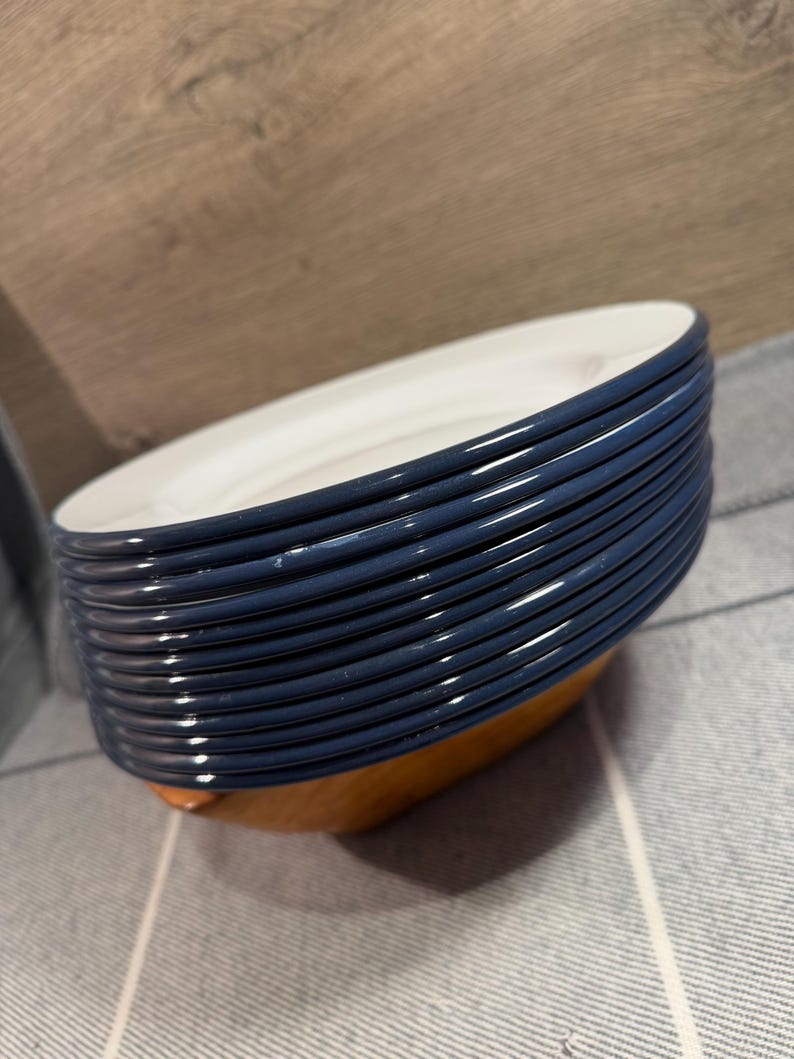 Pode incluir: Uma pilha de doze pratos redondos com uma borda azul-escura brilhante e um centro branco liso. Os pratos est&atilde;o dispostos numa pilha vertical, exibindo as bordas azuis uniformes e o design circular limpo do conjunto de lou&ccedil;as.
