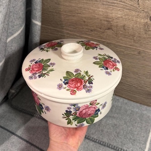 May include: A white ceramic lidded dish with a floral pattern of pink roses, purple flowers, and green leaves. The dish has a small handle on the lid, suitable for serving or storage.