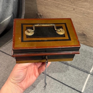 May include: An antique metal cash box with a key. The box is rectangular with a gold, black, and red color scheme. It has a brass handle and a keyhole with a key attached. The box is held by a person.