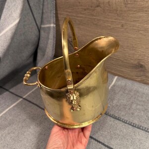 May include: A vintage brass coal scuttle with a handle and a spout. The scuttle has a lion's head detail on the front and a curved handle. The brass has a warm, golden color, and the scuttle appears to be well-preserved.