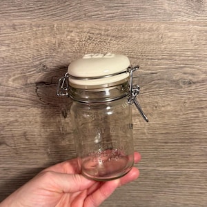 Pode incluir: Um frasco de vidro transparente com tampa de cerâmica creme e fecho de metal. O frasco tem o texto "Yorkshire England Est. 1900". O frasco é segurado em uma mão contra um fundo de madeira.