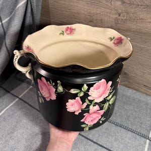Puede incluir: Jardinera de cerámica negra con diseño floral de rosas rosas. La jardinera tiene un borde festoneado con ribete dorado y dos asas. El interior es de color crema.