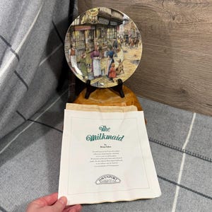 May include: A decorative plate depicting a street scene, displayed on a wooden stand. The plate shows a colourful illustration of people and buildings. A paper with the title "The Milkmaid" is in front of the plate.