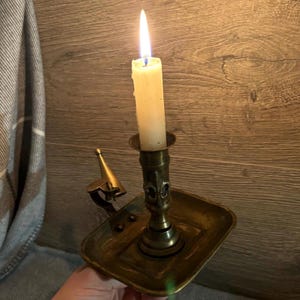 May include: A brass candle holder with a lit candle and a decorative design. The holder has a square base and a tall, ornate stem. The candle is white and is burning brightly.