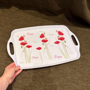 May include: A rectangular white serving tray with integrated side handles, featuring a repeating botanical print of red poppy flowers on green stems and the word Poppy printed in various sizes and shades of pink and green across the surface.