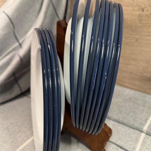 Pode incluir: Um conjunto de pratos de cer&acirc;mica branca com bordas azul-escuras, organizados em duas pilhas e mantidos na vertical por um suporte de madeira castanha. Os pratos apresentam um acabamento liso e brilhante nas bordas e um design circular.