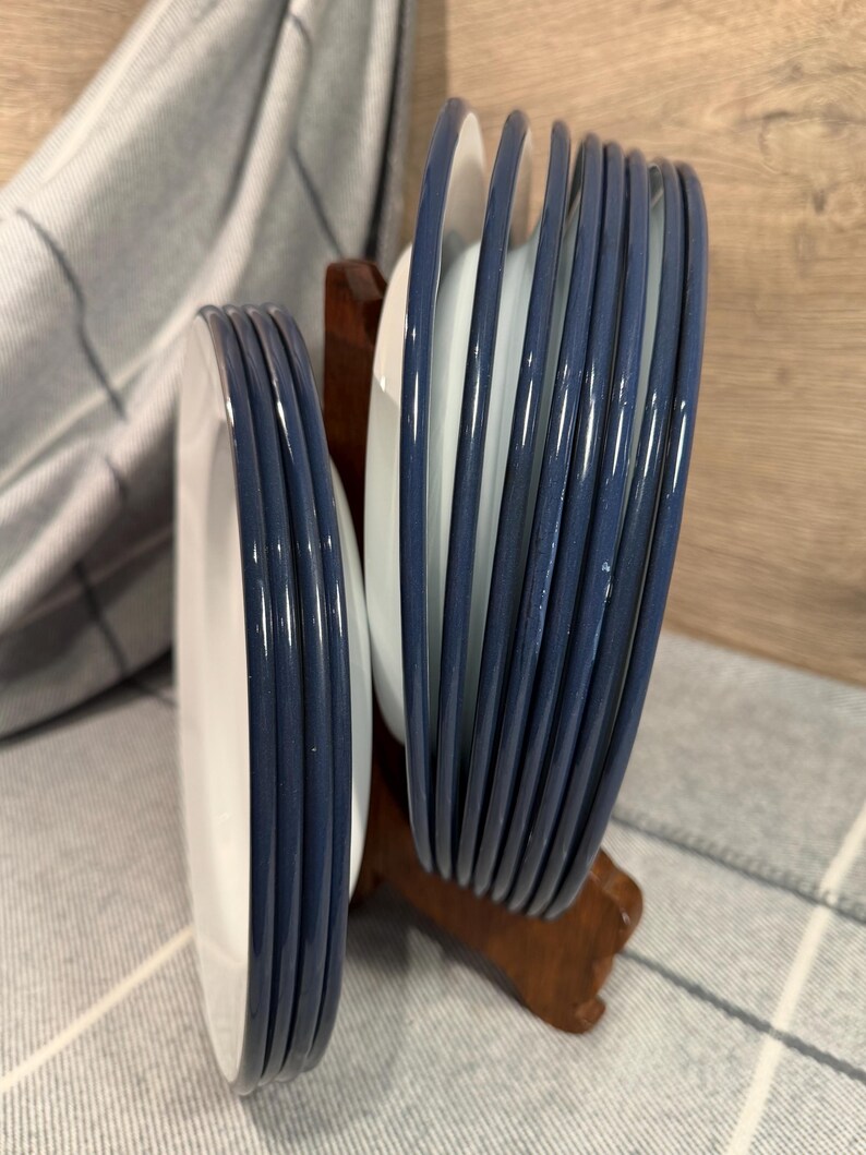 Pode incluir: Um conjunto de pratos de cer&acirc;mica branca com bordas azul-escuras, organizados em duas pilhas e mantidos na vertical por um suporte de madeira castanha. Os pratos apresentam um acabamento liso e brilhante nas bordas e um design circular.