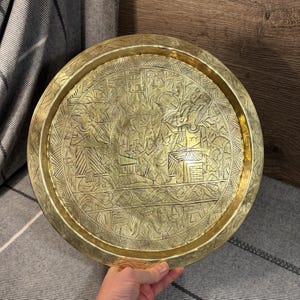 Puede incluir: Una bandeja redonda de latón con un diseño grabado intrincado que presenta figuras y símbolos. La bandeja tiene un borde elevado y un acabado pulido.