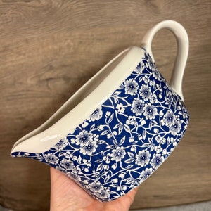 May include: A white ceramic gravy boat with a blue and white floral pattern. The boat has a wide, rounded base and a spout on one side. The handle is on the opposite side of the spout.