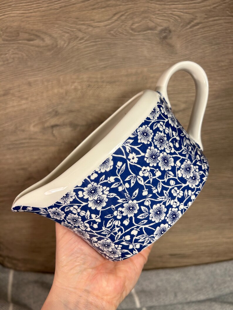 May include: A white ceramic gravy boat with a blue and white floral pattern. The boat has a wide, rounded base and a spout on one side. The handle is on the opposite side of the spout.