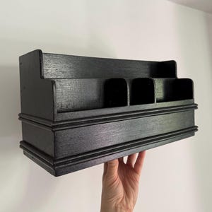 May include: A black wooden organiser with multiple compartments and a tiered design. The organiser has a rectangular shape with a decorative base. The compartments are of varying sizes. It is held by a hand against a white background.