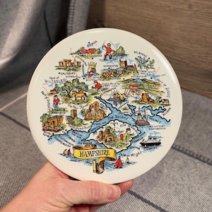 May include: A colourful ceramic plate featuring a map of Hampshire, England. The map highlights various towns and cities, including Winchester, Southampton, and Portsmouth. The plate is decorated with a blue and white border and the words "Hampshire" in a yellow banner.