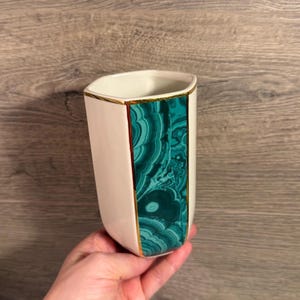 May include: A cylindrical ceramic tumbler featuring a smooth off-white finish and a vertical panel of green malachite pattern. The rim and the edges of the malachite inlay are accented with a thin, reflective gold-colored metallic trim.