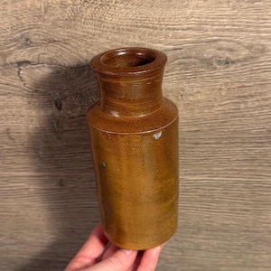 May include: A brown stoneware bottle with a cylindrical body and a slightly flared rim. The bottle has a smooth, glazed surface with some minor imperfections. The color is a warm, earthy brown, suggesting a vintage or antique piece. The bottle is held in a hand.