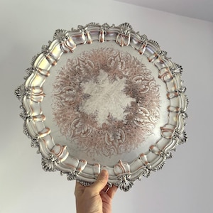 May include: Silver-toned decorative tray with an ornate, scalloped edge and a detailed floral design in the center. The tray has a vintage aesthetic, with a mix of silver and rose gold accents. The tray is held by a hand.