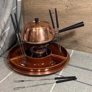 Puede incluir: Una olla de fondue de cobre con una base de cerámica marrón y seis tenedores de metal. La olla tiene una tapa y un quemador debajo. Los tenedores están dispuestos en un círculo alrededor de la olla.
