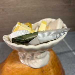 Op de afbeelding: Een witte, schelpvormige schaal van keramiek met een decoratieve basis, gevuld met kleine stukjes zeep en een klein pakje handcr&egrave;me.