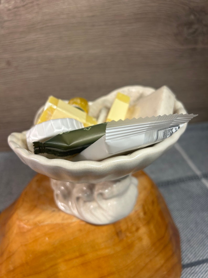 Op de afbeelding: Een witte, schelpvormige schaal van keramiek met een decoratieve basis, gevuld met kleine stukjes zeep en een klein pakje handcr&egrave;me.