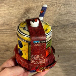 Könnte beinhalten: Eine farbenfrohe Keramikdose mit Deckel und kleinem Löffel. Die Dose hat ein rot-gelb-grünes Design mit verschiedenen Früchten und Gemüse. Der Text auf der Dose lautet "Pot et cuillère à confiture du grand marché".
