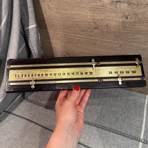 Puede incluir: Un dispositivo de medición negro y dorado antiguo con números del 1 al 20 y del 20 al 100. El dispositivo tiene una escala de color latón y lo sostiene una persona con esmalte de uñas rojo.