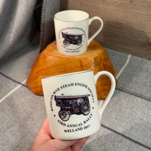 May include: Two white ceramic mugs with a blue rim and handle. Each mug features a vintage steam engine illustration and text that reads "ROSS-ON-WYE STEAM ENGINE SOCIETY 53RD ANNUAL RALLY WELLAND 2017".