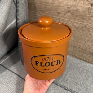 Puede incluir: Un bote de harina de cerámica naranja con tapa y pomo. La palabra "FLOUR" está impresa en negro sobre una etiqueta ovalada. El bote está en la mano de una persona. Ideal para la cocina.