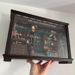 May include: A framed print of "The Old Curiosity Shop" in a dark brown wooden frame. The print shows a detailed interior scene of a shop filled with books, tools, and decorative objects. The shop's interior is painted in shades of green and brown.