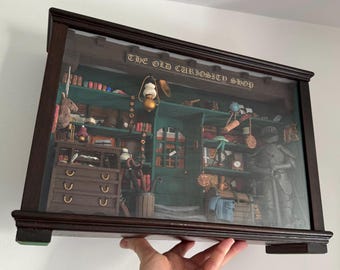 May include: A framed print of "The Old Curiosity Shop" with a dark brown wooden frame. The print depicts a detailed interior scene of a shop filled with various items, including books, tools, and decorative objects. The shop's interior is painted in shades of green and brown.
