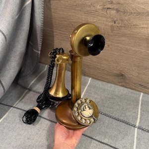 Puede incluir: Un teléfono de columna de latón de estilo vintage con un dial giratorio que presenta un gráfico central de una persona y el texto "CONVERSATION PIECES". El dispositivo incluye un auricular desmontable conectado por un cable negro en espiral.