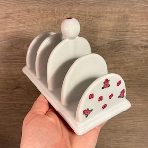 Puede incluir: Tostador de cerámica blanco con cuatro ranuras y una pequeña perilla en la parte superior. El tostador tiene un diseño floral con rosas rosas en la parte inferior.