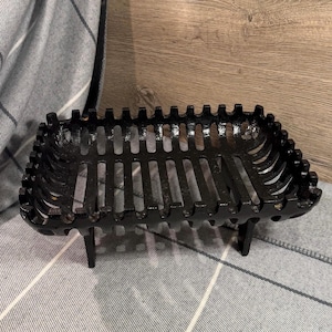 May include: A rectangular black cast iron fireplace grate featuring a slatted base and vertical side bars with a notched top edge. The grate is supported by four short, sturdy legs, providing a raised structure for holding firewood securely.
