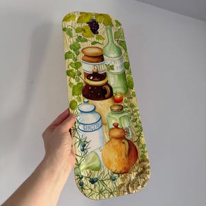 May include: A rectangular serving tray with a painted design of kitchen items. The tray features a light wood grain background with illustrations of jars, bottles, and a bunch of grapes. The tray is held by a person's hand.