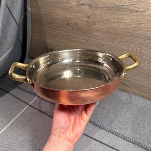 May include: A round cooking pan with a polished copper exterior and a shiny silver-toned interior. The pan features two textured, gold-toned metal handles attached to the sides with rivets, providing a sturdy grip for this kitchen cookware item.