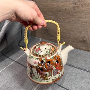 Puede incluir: Una tetera de cerámica blanca con un diseño de inspiración asiática. Tiene un asa de bambú y una tapa decorada con flores y pájaros. El cuerpo de la tetera muestra figuras en un jardín.