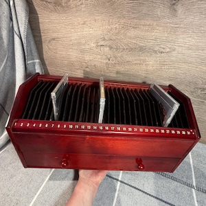 Puede incluir: Un mueble de almacenamiento de CD de madera de color rojo oscuro. Cuenta con ranuras numeradas para CD, con un cajón debajo. Varios CD son visibles en las ranuras. Los números del 1 al 31 están impresos en la parte delantera.