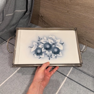 Op de afbeelding: Een rechthoekig dienblad met een zilverkleurig metalen frame en twee handgrepen aan de zijkanten. Het midden heeft een witte keramische basis versierd met een blauwe illustratie van drie ronde vruchten omringd door bladeren op een witte achtergrond.
