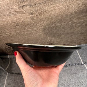 May include: A black ceramic bowl with a white rim is held in a hand. The bowl has a glossy finish and a simple, classic design. The background is a wooden surface.