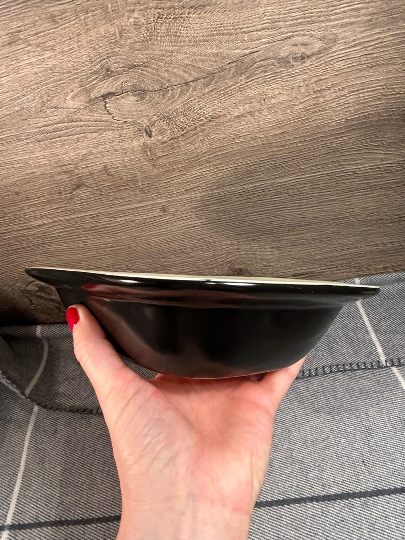 May include: A black ceramic bowl with a white rim is held in a hand. The bowl has a glossy finish and a simple, classic design. The background is a wooden surface.